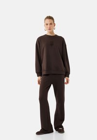 Sudadera de algodón marrón con hombros caídos y texto bordado, combinada con pantalones marrones a juego. La sudadera tiene un corte holgado y puños acanalados.
