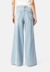 Személy világoskék, bőszárú farmernadrágban áll, amelyen hímzett "ER" látható a jobb hátsó zsebnél, egy sima háttér előtt.