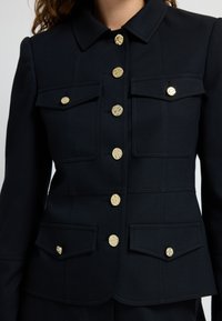 Czarna dopasowana marynarka z kołnierzykiem, wyposażona w złote guziki i dwie przednie kieszenie z klapkami. Teksturowany materiał.