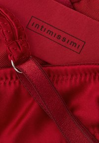 Lencería roja con tirantes intrincados, superficie de tela suave y una etiqueta de marca que muestra "intimissimi" en letras negras.
