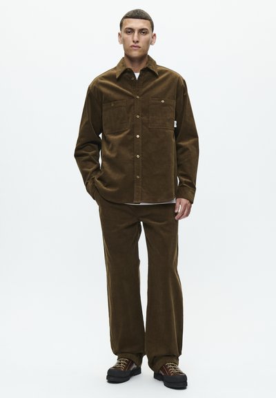 Veste en velours côtelé marron avec deux poches avant, pantalon en velours côtelé assorti et chaussures foncées. Design simple avec texture verticale.