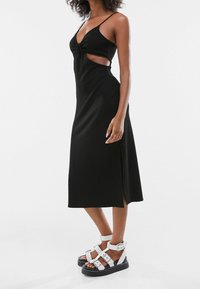 Vestido midi negro de canalé con un detalle frontal retorcido, cortes laterales y tirantes finos. Combinado con sandalias de plataforma blancas con hebillas.