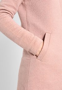 Vaaleanpunainen teksturoitu mekko, jossa on ribbitetty kuvio, pitkät hihat ja sivutaskut. Valmistettu pehmeästä kankaasta mukavuutta varten.