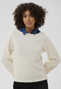 Pull en maille texturée écru avec un large col, des manches longues et une coupe décontractée, superposé à un col de chemise en denim foncé.