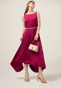 Fuksia ilmanhihaine mekko, jossa on korkea-alahaem, jota korostaa hopeinen vyö. Mallilla on kädessään pieni beige laukku ja hän on pukenut päälleen vaaleat sandaalit.