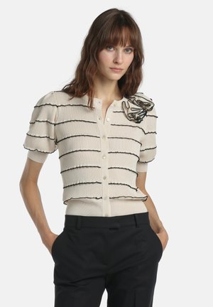 Barna hajú nő, bézs rövid ujjú, gombos felsőben fekete csíkokkal és virágdísszel, fekete nadrággal, kezét zsebbe dugva.