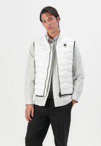 White quilted puffer vest with a high collar. Features black zipper and accents, worn over a gray ribbed sweater and striped button-up shirt.