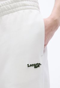 Χέρι στην τσέπη παντελονιού με άσπλη ελαστική μέση και εμφανές πράσινο λογότυπο Lacoste στο αριστερό μηρό, σε λευκό φόντο.