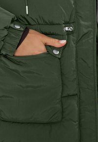 Chaqueta acolchada verde con una textura suave y mate. Presenta un bolsillo lateral asegurado con botones a presión plateados y mangas fruncidas para un ajuste ceñido.