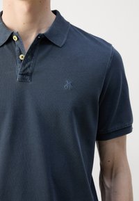 Homme portant un polo bleu foncé avec des boutons jaunes et un petit logo MOP brodé sur la poitrine gauche.