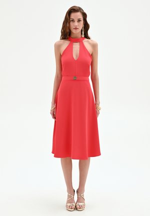 Femme portant une robe midi rouge sans manches avec un col licou, une découpe en forme de goutte, une ceinture assortie, des sandales dorées et des boucles d'oreilles remarquables, debout, le dos droit.