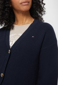 Marinblå texturerad cardigan med djup V-halsringning, träknappar och en liten logotyp på bröstet. Bärs över en ljusfärgad skjorta.