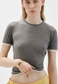 Camiseta gris de manga corta con ribete blanco y textura acanalada. El modelo lleva una pulsera de oro en una muñeca y una falda amarilla.