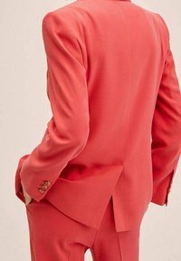 Korallefarbener Anzug, bestehend aus einem taillierten Blazer mit einem Schlitz und passenden Hosen. Der Stoff wirkt weich mit minimaler Textur; Knöpfe sind sichtbar.