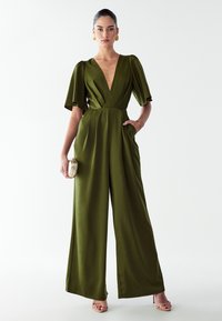 Ženska stoji oblečena v olivno zeleni široki kombinezon s napihnjenimi rokavi in globokim V-izrezom, v roki drži majhno zlat kluč in nosi zlate uhane.