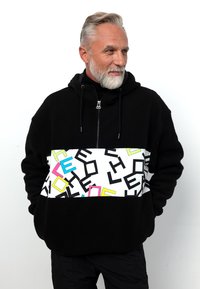 Schwarze Fleece-Hoodie mit einem weißen Abschnitt, der farbenfrohe geometrische Muster in Rosa, Blau und Gelb aufweist. Mit Reißverschluss am Kragen, lockere Passform.