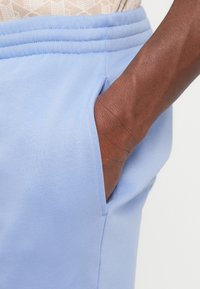 Mano nella tasca dei pantaloni della tuta azzurri con banda elastica a costine in vita, camicia beige parzialmente visibile sopra i pantaloni.