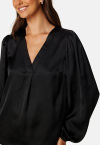 Blusa nera di satin con scollatura a V, maniche a palloncino e vestibilità morbida. La trama liscia e la leggera lucentezza sono caratteristiche predominanti.