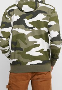 Maskáčová mikina s kapucí v olivově zelené, černé a bílé barvě. Materiál z bavlněné směsi s kapucí ve stylu klokaní kapsy a žebrovanými manžetami.