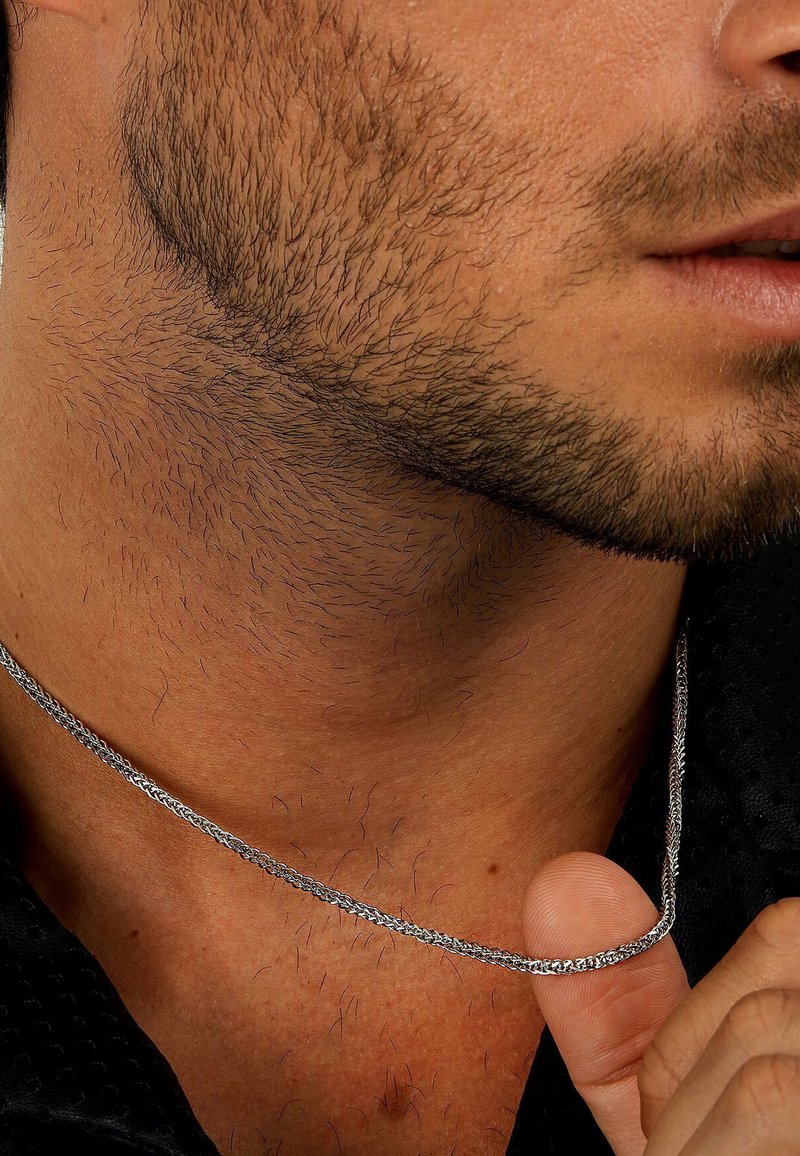 Silberne Halskette in einem geflochtenen Design, die auf der Haut ruht, hervorgehoben durch Gesichtshaare und einen Finger, der die Kette sanft hält.