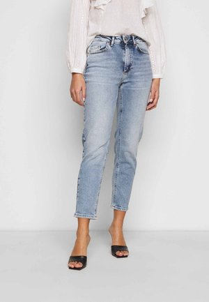 Frau trägt hellblaue High-Waist-Jeans mit geradem Bein, weiße Langarmbluse und schwarze Sandaletten mit offenem Zehenbereich, steht vor weißem Hintergrund.