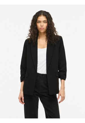 Ženska s kodrastimi lasmi, oblečena v črni blazer čez belo majico in črne hlače, stoji pred enostavnim belem ozadjem.