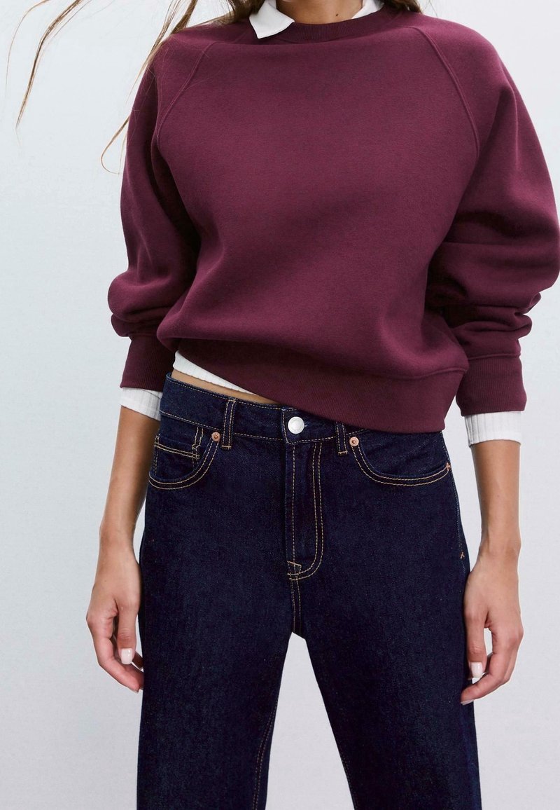 Femme portant un sweat-shirt bordeaux sur une chemise blanche à col, associée à un jean taille haute bleu foncé, debout devant un fond uni.