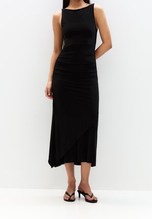Mujer con vestido midi asimétrico negro sin mangas y sandalias negras de tacón alto, de pie sobre un fondo claro.