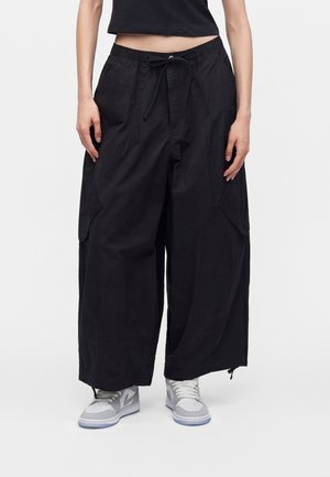 Person iført løse sorte cargo bukser med brede ben, grå og hvide sneakers samt en sort crop top mod en ensfarvet baggrund.