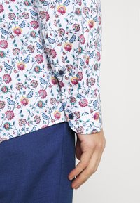 Chemise blanche à motif floral multicolore. Tissu doux, manches longues avec poignets à boutons, associée à un pantalon bleu foncé. Design décontracté.