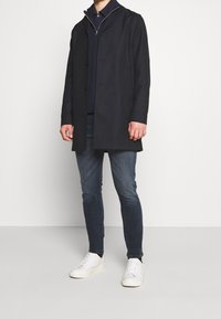 Marinblå kappa med krage, två knappar och sidofickor, tillsammans med en mörk tröja och ljusa jeans. Avslutat med vita sneakers.