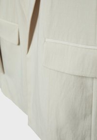 Gros plan sur un tissu beige présentant deux poches à rabat rectangulaires sur une veste ou un manteau avec des détails de coutures visibles.
