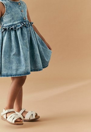 Enfant portant une robe en jean sans manches bleue avec des boutons et des sandales blanches, tenant l'ourlet de la robe contre un fond beige.