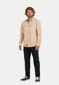 Beige Hemd mit Knopfleiste und zwei Brusttaschen, kombiniert mit schwarzen Hosen und grünen Sneakers. Der Stoff hat eine strukturierte Oberfläche.