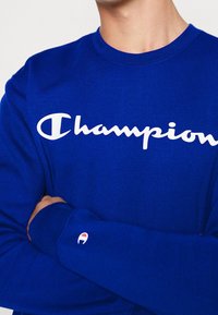 Modrá bavlněná mikina se bílým logem "Champion" přes prsa. Má žebrovaný výstřih a malé logo na rukávu.