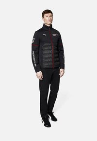 Czarna puchowa kurtka z czerwonymi akcentami, z zapięciem na zamek i bocznymi panelami. Noszona z czarnymi dżinsami i butami, na białym tle.