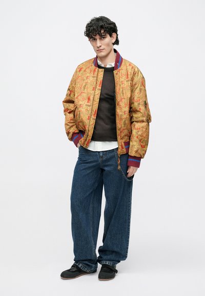 Zlatá bomber bunda so vzorom, s rebrovan�ým golierom a manžetami, v kombinácii s tmavým svetrom, bielou košeľou a voľnými džínsami. Čierne topánky.