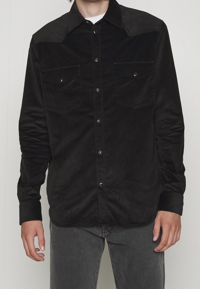 Camicia di velluto a coste nera con chiusura a bottoni frontale, due tasche sul petto e dettaglio del giogo appuntito. Presenta maniche lunghe con polsi abbottonati.