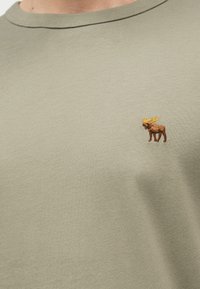 Ανοιχτό πράσινο πουκάμισο με μικρό κεντητό καφέ ελάφι με κίτρινα κέρατα στην αριστερή πλευρά του στήθους.