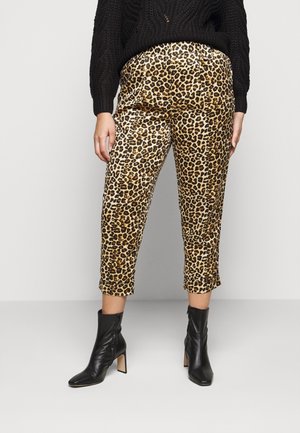 Femme portant un pantalon court imprimé léopard, un pull noir et des bottines à talons noires, debout devant un fond clair uni.