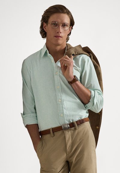 Light green striped button-up shirt with rolled sleeves, paired with beige pants and a brown belt. Holding a brown houndstooth blazer.