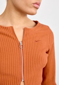 Top naranja de canalé con escote redondo, media cremallera con tirador circular y un pequeño logo bordado en el pecho.