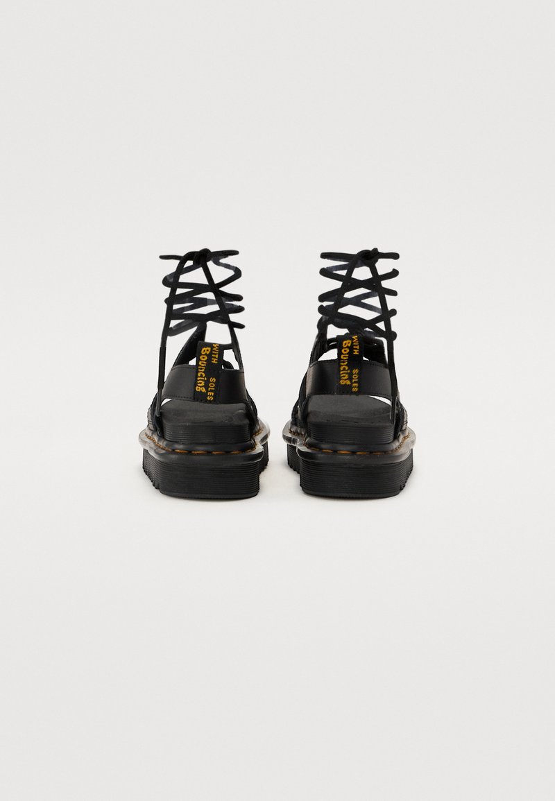 Black strappy sandals feature a chunky platform sole, zigzag laces, and contrasting yellow branding on the heel. Smooth leather texture.