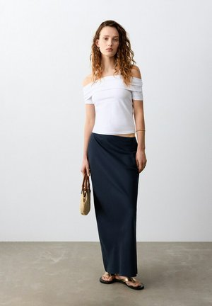 Jeune femme aux cheveux bouclés portant un haut blanc à épaules dénudées, une longue jupe noire, tenant un sac tissé, debout sur un sol en béton devant un mur blanc.