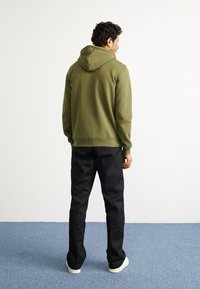 Sweatshirt de um verde oliva com bolso frontal e cordão, combinado com calças pretas e tênis brancos. Tecido macio e texturizado.