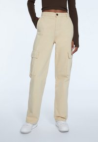 Osoba w beżowych spodniach cargo z bocznymi kieszeniami, brązowej bluzie z długim rękawem i białych trampkach, stoi z rękami w kieszeniach.