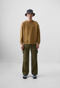 Hnedá mikina s okrúhlym výstrihom, zelené cargo nohavice so bočnými vreckami a šedý vedierkový klobúk. Čierne a modré tenisky dopĺňajú outfit.
