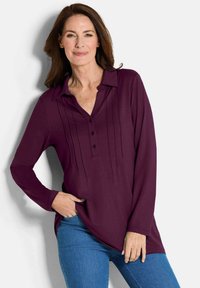 Langärmlige Bluse in Pflaumenfarbe mit Kragen, Knopfleiste und vertikalen Falten. Kombiniert mit blauen Jeans steht das Modell vor einem neutralen Hintergrund.