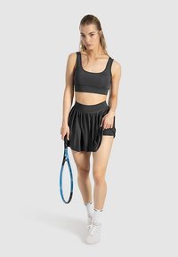 Sort atletisk crop top og plisseret nederdel, holder en tennisketcher. Stoffet virker glat. Sokker og sneakers er hvide. Simpelt design.