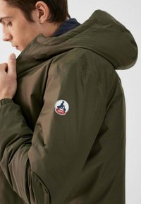 Veste légère vert olive avec capuche, poignets élastiques et un patch rond du logo JOTT sur la manche gauche. Texture lisse et design minimaliste.